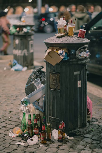 A black cylindrical rubbish bin made of metal is positioned on a cobblestone pavement outdoors, filled with discarded paper and various waste items. The lid of the bin is open, with several empty glass bottles, crumpled paper, and small food containers visible around and on top of it. Several bottles, some with labels, are scattered on the ground in front of the bin, with a few crumpled napkins and pieces of trash nearby. The surrounding area includes a background of parked cars, a grey rubbish bin, and blurry pedestrians, indicating a busy urban environment. The lighting suggests daytime, with natural light illuminating the scene, emphasizing the clutter and litter that suggest improper disposal or overfilled waste receptacles, typical of areas where private rubbish collection or citizen waste management issues occur. This scene aligns with topics related to rubbish removal services and the importance of maintaining clean public spaces, highlighting the role of waste collection providers such as Rubbish Collection Dulwich in managing urban waste in East Dulwich SE22 and surrounding areas.
