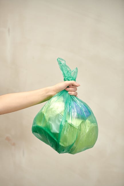 A person's arm extends into the frame from the left, holding a green and blue plastic rubbish bag by its twisted top. The bag appears full and is bulging, with visible creases and a slightly glossy surface. The background is plain and neutral, with a soft beige tone, giving a minimalistic environment that emphasizes the bag and the hand. The scene suggests an informal waste disposal or rubbish collection context, with the bag likely containing household waste or rubbish for collection. The lighting is even and natural, highlighting the colors and textures of the plastic bag without shadows or reflections. This image exemplifies typical practices of on-site rubbish handling in domestic or small-scale waste management settings, aligning with independent disposal and waste collection activities undertaken by services like Rubbish Collection Dulwich.