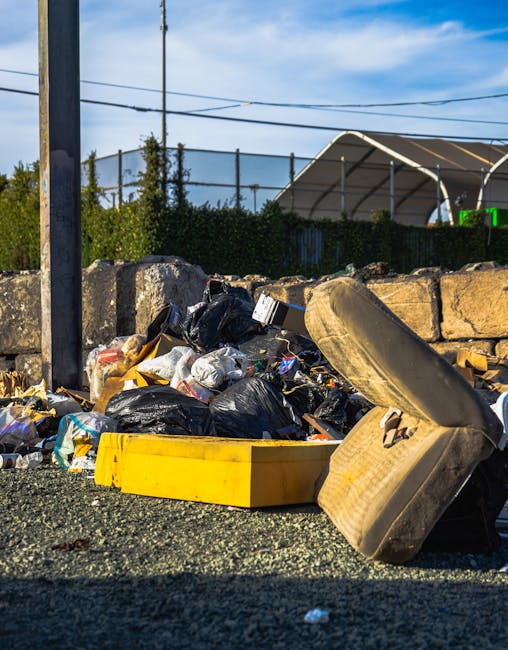 A close-up view of a pile of mixed waste materials on a gravel surface, including black and clear plastic rubbish bags, some partially torn open revealing packaging and debris, alongside an old, dirty beige car seat with fabric upholstery and visible wear, leaning against a low stone wall constructed of irregularly shaped stones. Behind the waste, there is a green hedgerow and a large sports net or enclosure with a curved roof structure, supported by metal poles, suggesting an outdoor sports facility. Overhead, power lines and a wooden utility pole are visible under a partly cloudy sky with blue and white tones. The scene appears to depict an informal rubbish accumulation at the edge of a property or public area, possibly related to private waste disposal rather than official collection, consistent with independent rubbish removal services such as those provided by Rubbish Collection Dulwich.