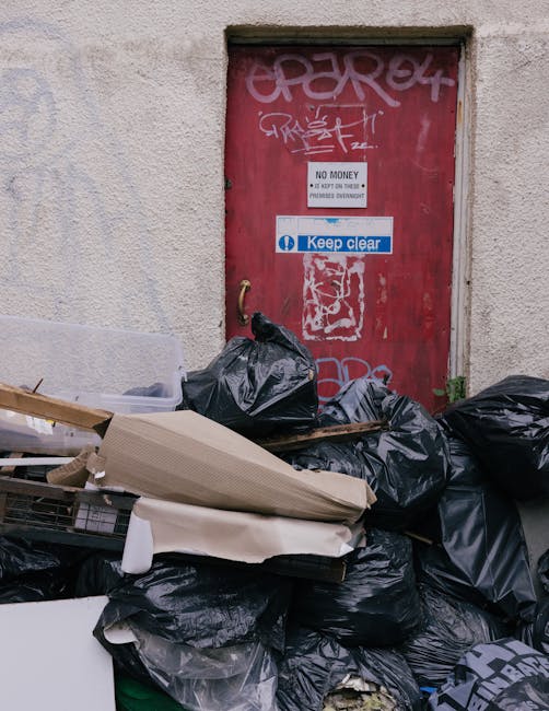 A large pile of black rubbish bags filled with waste sits in front of a weathered red metal door attached to a textured off-white wall. The bags are stacked in a disorderly manner, with some leaning against each other and others placed on top of cardboard and loose debris. The cardboard appears partially flattened, with torn edges and creases, and is positioned across the top of the bags. The red door features various graffiti markings in white and pink spray paint, with some overlapping. Several signs are affixed to the door: a white sign stating 'NO MONEY' and 'KEEP CLEAR', and a blue and white sticker that reads 'Keep clear'. The door has a brass handle on the left side and shows signs of rust and peeling paint around the edges. The surface surrounding the door is rough textured, indicating an exterior wall. This scene likely depicts an area where independent waste collection or on-site rubbish disposal occurs, highlighting the importance of professional waste removal services like those offered by Rubbish Collection Dulwich to manage excess waste efficiently and maintain environmental standards.