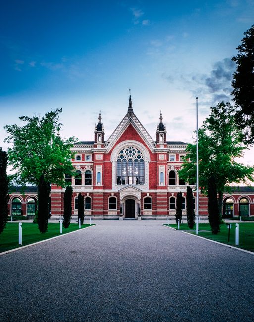The image depicts the facade of a historic red brick building with intricate architectural details, including large arched windows, decorative stonework, and a prominent central entrance. The structure features a steeply pitched roof with decorative turrets and spires, set against a partly cloudy sky during daylight hours. Flanking the building are evenly spaced, mature green trees with lush foliage, and a paved pathway leads directly to the entrance, bordered by short white posts. The surrounding area appears to be a well-maintained garden or plaza with neatly trimmed grass and a few visible street lamps, suggesting a public or institutional setting. The scene captures a clean, organized exterior typical of a cultural or historic site, with no visible rubbish or debris, reflecting a maintained environment. Although the location is not specified, the scene echoes the kind of well-preserved private grounds or entrance areas associated with significant cultural landmarks, which might require occasional external waste handling or rubbish removal services, such as those provided by Rubbish Collection Dulwich for maintaining pristine external environments outside heritage sites or public buildings.
