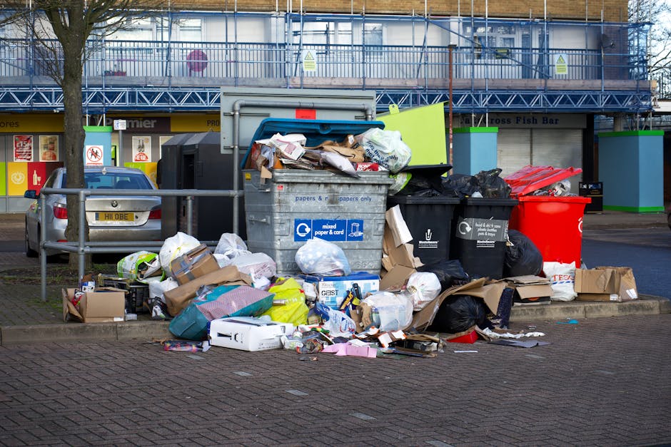 A black, cast-iron public trash bin situated on a cobblestone pavement in an urban environment, with various rubbish items surrounding and atop it. The bin has a domed lid with a circular opening, and is covered with litter, including several glass bottles, some crumpled paper, and food packaging. On top of the bin, there are disposable coffee cups, a paper food container, and a small cardboard box, indicating nearby food consumption. Additional rubbish, such as plastic bags filled with trash, is piled at the base and nearby, with some waste spilling onto the pavement. In the background, a cluttered street scene with blurred figures, parked cars, and scattered urban debris is visible, suggesting an area with frequent foot traffic and informal waste disposal, which Rubbish Collection Dulwich occasionally manages through independent collection and on-site clearance services. The overall atmosphere reflects typical urban littering in a busy public space, highlighting the importance of waste management and proper disposal practices in city environments.