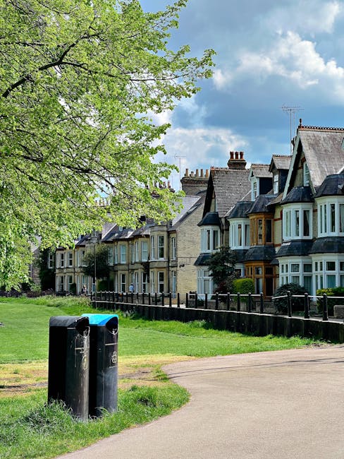 A pair of black rubbish bins positioned on a grassy verge adjacent to a paved footpath in front of a row of historic terraced houses with bay windows, pitched slate roofs, and brick chimneys. The bins are situated near a low wooden fence that separates the pavement from the garden frontages of the houses. A large leafy tree with bright green foliage extends its branches over the scene, providing shade and adding natural greenery to the environment. The sky above is partly cloudy with patches of blue, indicating a bright day. The setting appears to be a residential area with typical architecture and outdoor space suitable for private rubbish collection or on-site waste management, similar to what might be handled by a local rubbish removal service like Rubbish Collection Dulwich. The scene emphasizes a tidy, well-maintained neighborhood with clear access for waste disposal, reflecting standard practices for suburban rubbish management.