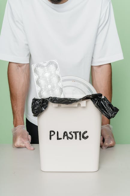 A person wearing a white T-shirt and transparent gloves stands behind a white plastic bin labeled 'PLASTIC'. The bin is partially open with a black plastic bag lining its interior, which is visible at the top. A clear plastic packaging tray with multiple compartments rests on the edge of the bin, positioned to the left. The background features a plain light green wall, and the scene is evenly lit, emphasizing the focus on waste materials and the disposal process. This setup suggests an environment related to waste collection or recycling, typical of private or independent rubbish removal services like those offered by Rubbish Collection Dulwich, focusing on the proper separation and disposal of plastics within a waste management context.